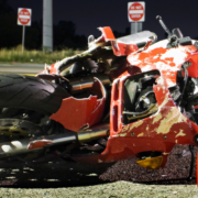 A dynamic and professional image designed for Alabama riders. On the right, a motorcycle stands parked on a coastal highway overlooking the Mobile Bay bridge at sunset. On the left, bold, high-contrast text overlays a darkened background: "What Is the Average Settlement for a Motorcycle Crash in Alabama?" Below, the Quin Baker Law logo is visible alongside a "Free Case Evaluation" button.