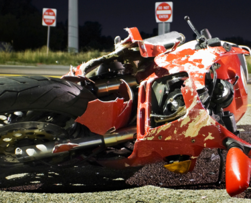 A dynamic and professional image designed for Alabama riders. On the right, a motorcycle stands parked on a coastal highway overlooking the Mobile Bay bridge at sunset. On the left, bold, high-contrast text overlays a darkened background: "What Is the Average Settlement for a Motorcycle Crash in Alabama?" Below, the Quin Baker Law logo is visible alongside a "Free Case Evaluation" button.