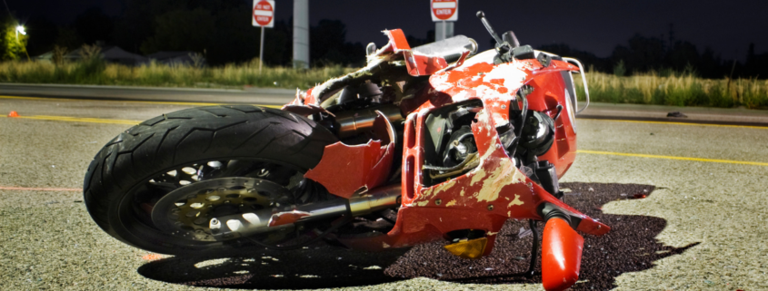A dynamic and professional image designed for Alabama riders. On the right, a motorcycle stands parked on a coastal highway overlooking the Mobile Bay bridge at sunset. On the left, bold, high-contrast text overlays a darkened background: "What Is the Average Settlement for a Motorcycle Crash in Alabama?" Below, the Quin Baker Law logo is visible alongside a "Free Case Evaluation" button.